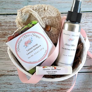 May include: A gift basket with a variety of Ramblin' Rose Soaps products. The basket contains a bottle of body mist, a bar of tallow soap, and a lip balm. The products are packaged in a white basket with a pink ribbon and a beige washcloth.