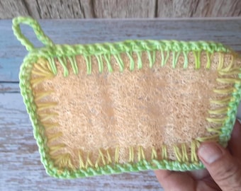 May include: A rectangular loofah sponge featuring a textured, light tan fibrous center. The edges are reinforced with a bright lime green crocheted border and decorative yellow stitching, including a small loop at the top corner for hanging.