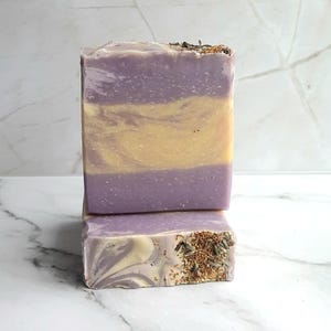 May include: Two rectangular bars of handmade soap. The top bar has layers of lavender and yellow, with dried herbs on top. The bottom bar has a swirl pattern with lavender and white, and is also topped with dried herbs. The soap is displayed on a marble surface.
