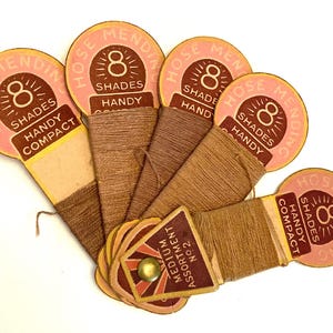 Op de afbeelding: Een verzameling vintage garenspoelen voor het repareren van kousen. De spoelen zijn in een waaier gerangschikt en tonen verschillende bruine tinten. Elke spoel heeft een roze en bruin label met de tekst "8 Shades Handy Compact".