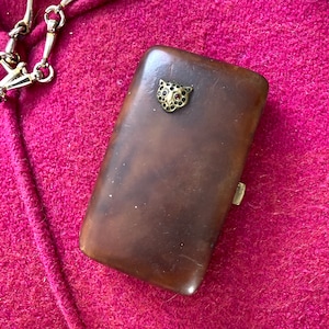 Puede incluir: Un estuche de cigarrillos de cuero marrón con un diseño de cabeza de leopardo en metal dorado. El leopardo tiene un ojo de piedra roja.
