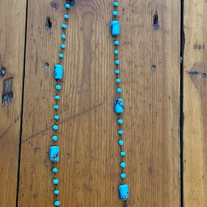 May include: A long turquoise blue necklace with rectangular beads on a brown wooden background.