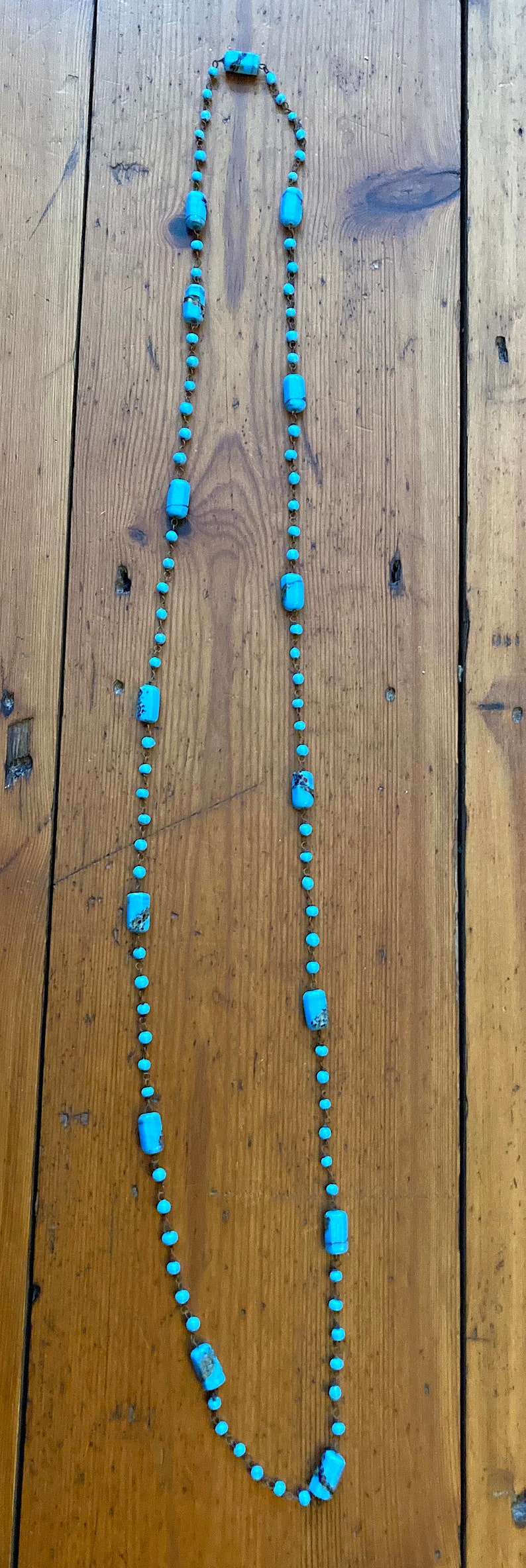 May include: A long turquoise blue necklace with rectangular beads on a brown wooden background.