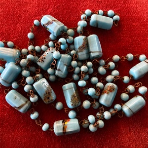 May include: A long necklace made of light blue glass beads with brown and gold flecks. The beads are rectangular and are strung on a thin copper chain.