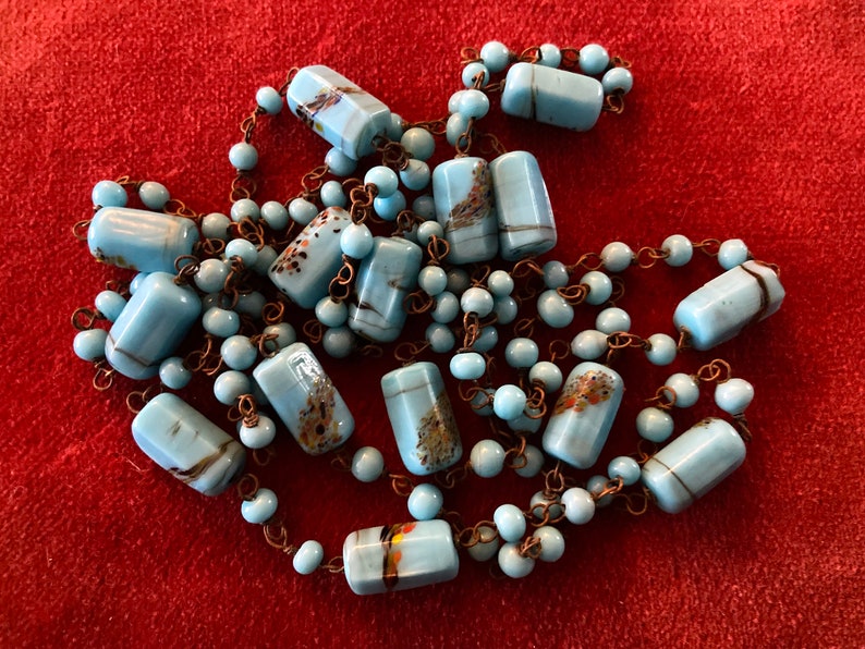 May include: A long necklace made of light blue glass beads with brown and gold flecks. The beads are rectangular and are strung on a thin copper chain.