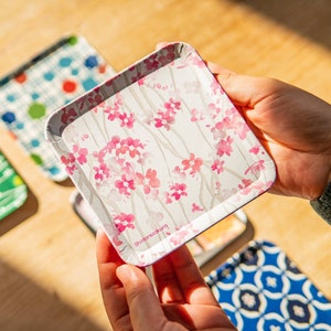 以下が含まれることがあります： 異なる花柄と幾何学模様の小さな正方形のトレイが5つセットになっています。トレイは金属製で、白い背景です。中央のトレイは手で持たれており、ピンクの花柄と右下に「shizukazuki」という文字が書かれています。