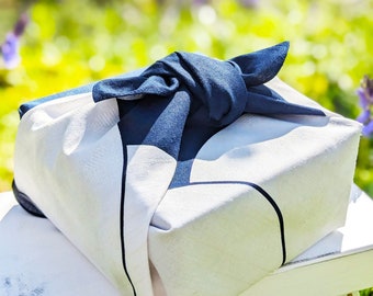 May include: A white gift box wrapped in a blue fabric with a knot on top. The box is sitting on a white table with a blurred green background.