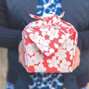 Puede incluir: Una caja de regalo cuadrada envuelta en tela roja con un estampado floral blanco. La caja está atada con un nudo de tela a juego. La tela tiene un patrón repetido de flores blancas sobre un fondo rojo.