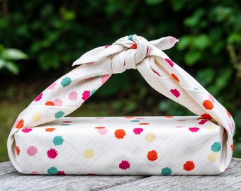May include: A white fabric gift wrap with a colorful polka dot pattern is tied around a rectangular box. The fabric is tied in a knot at the top of the box.