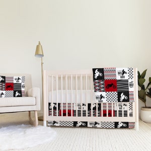 May include: A white crib with a red, black, and white patterned crib bedding set. The bedding features a checkered pattern, a wave pattern, and a dirt bike graphic. The crib is next to a white rocking chair with a red, black, and white patterned pillow. A red, black, and white patterned curtain is in the background.