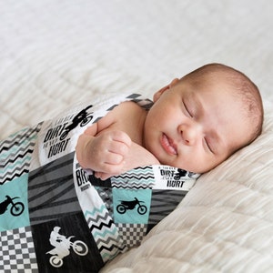 May include: A baby swaddled in a blue and gray baby blanket with a motocross theme. The blanket features a checkered pattern, a chevron pattern, and a wood grain pattern. The blanket also features the text "A little dirt never hurt".