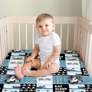 Op de afbeelding: Een baby zit in een wit ledikant met een blauw en zwart gepatroneerd hoeslaken. Het hoeslaken heeft een patchwork-design met afbeeldingen van crossmotoren en de tekst "Eat Sleep Ride".