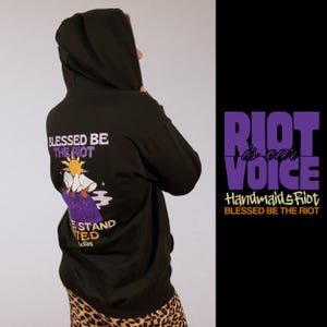 Pode incluir: Moletom preto com capuz com o texto "BLESSED BE THE RIOT" e um gr&aacute;fico nas costas. O design inclui um sol, uma figura em vestes roxas e as palavras "WE STAND UNITED". O lado direito apresenta o texto "RIOT is our VOICE".