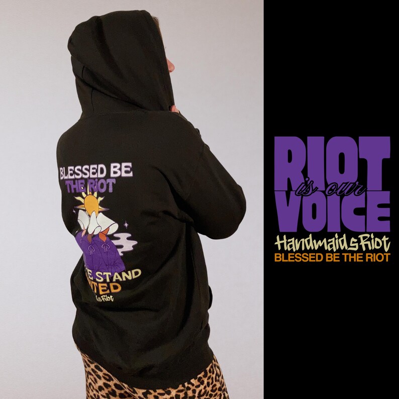 Pode incluir: Moletom preto com capuz com o texto "BLESSED BE THE RIOT" e um gr&aacute;fico nas costas. O design inclui um sol, uma figura em vestes roxas e as palavras "WE STAND UNITED". O lado direito apresenta o texto "RIOT is our VOICE".