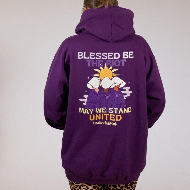 Pode incluir: Um moletom roxo com capuz com o texto "BLESSED BE THE RIOT" acima de um gr&aacute;fico de tr&ecirc;s figuras com capuzes brancos e o texto "MAY WE STAND UNITED". O moletom tem capuz e mangas compridas.