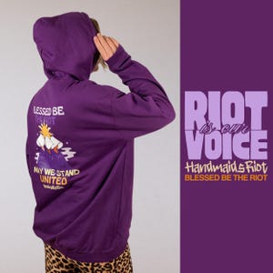 Pode incluir: Um moletom roxo com a frase "BLESSED BE THE RIOT" e "RIOT is our VOICE" impressas. A parte de tr&aacute;s do moletom apresenta uma ilustra&ccedil;&atilde;o com as palavras "MAY WE STAND UNITED".
