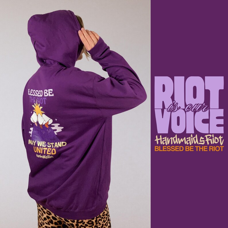 Pode incluir: Um moletom roxo com a frase "BLESSED BE THE RIOT" e "RIOT is our VOICE" impressas. A parte de tr&aacute;s do moletom apresenta uma ilustra&ccedil;&atilde;o com as palavras "MAY WE STAND UNITED".