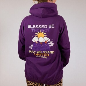 Pode incluir: Um moletom roxo com o texto "Blessed Be The Riot" e "May We Stand United" em branco e amarelo. O gr&aacute;fico apresenta tr&ecirc;s figuras com capuzes brancos e t&uacute;nicas roxas, com um sol ao fundo. O moletom &eacute; de cor roxa s&oacute;lida.