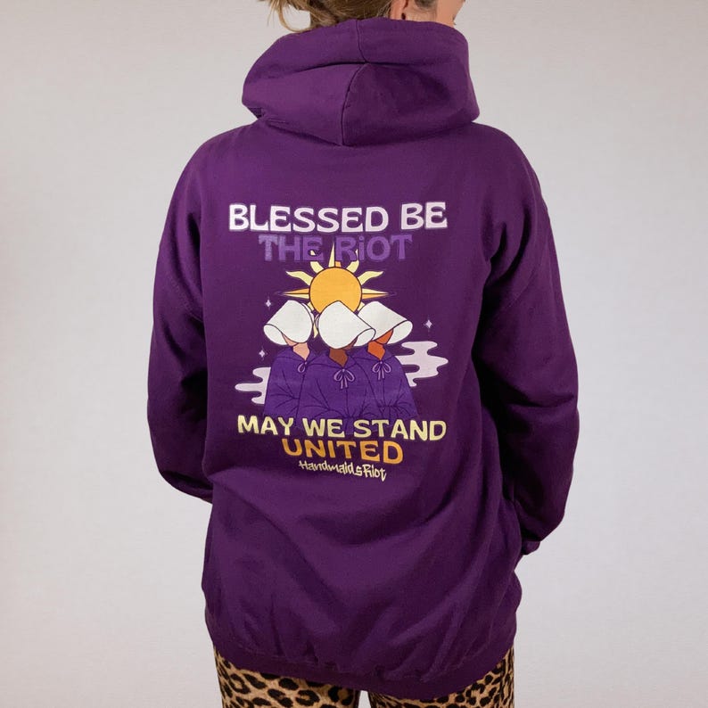 Pode incluir: Um moletom roxo com o texto "Blessed Be The Riot" e "May We Stand United" em branco e amarelo. O gr&aacute;fico apresenta tr&ecirc;s figuras com capuzes brancos e t&uacute;nicas roxas, com um sol ao fundo. O moletom &eacute; de cor roxa s&oacute;lida.