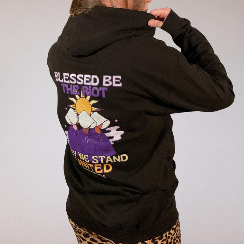 Pode incluir: Moletom preto com a frase "BLESSED BE THE RIOT" acima de uma ilustra&ccedil;&atilde;o de tr&ecirc;s figuras em vestes roxas e toucas brancas. A frase "WHY WE STAND UNITED" est&aacute; abaixo. O moletom &eacute; usado por uma pessoa.
