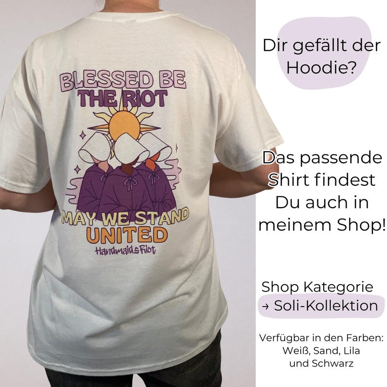 Pode incluir: Camiseta creme com um gr&aacute;fico nas costas com o texto "BLESSED BE THE RIOT" e "MAY WE STAND UNITED". O design inclui figuras com capas roxas e toucas brancas, com um sol. Parte da Soli-Kollektion. Dispon&iacute;vel em branco, areia, lil&aacute;s e preto.