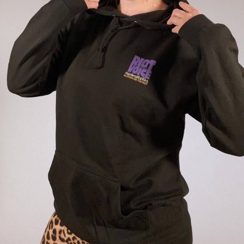 Pode incluir: Moletom preto com capuz, bolso frontal e cord&atilde;o. O texto roxo "RIOT VOICE" &eacute; impresso na parte superior esquerda do peito, com o texto "Handmaids & Riot" abaixo em dourado.