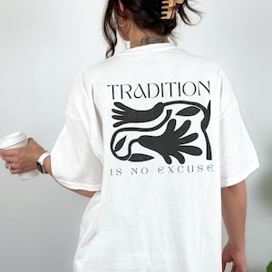 Może przedstawiać: Biała koszulka z czarnym nadrukiem graficznym na plecach. Projekt zawiera słowo "TRADITION" nad stylizowaną ilustracją i frazę "IS NO EXCUSE" poniżej. Koszulka wykonana jest z miękkiego materiału.