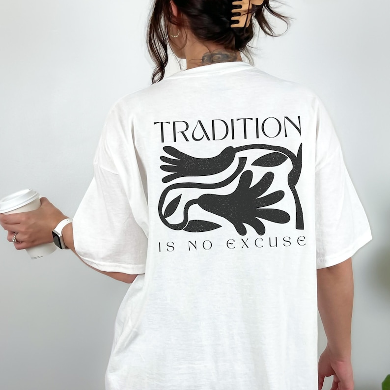 Może przedstawiać: Biała koszulka z czarnym nadrukiem graficznym na plecach. Projekt zawiera słowo "TRADITION" nad stylizowaną ilustracją i frazę "IS NO EXCUSE" poniżej. Koszulka wykonana jest z miękkiego materiału.