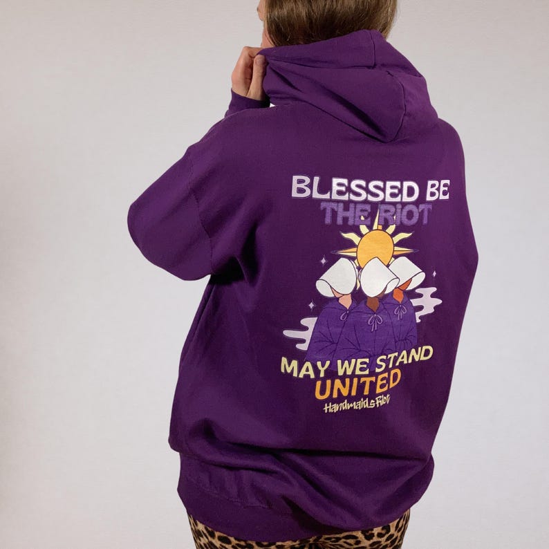 Pode incluir: Um moletom roxo com o texto "BLESSED BE THE RIOT" e "MAY WE STAND UNITED" em branco e amarelo. O gr&aacute;fico apresenta tr&ecirc;s figuras com toucas brancas e capas roxas, com um sol ao fundo. Handmade fibers.