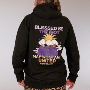 Pode incluir: Moletom preto com a frase "Blessed Be The Riot" e "May We Stand United" em roxo e amarelo. O gr&aacute;fico apresenta tr&ecirc;s figuras com toucas brancas e vestes roxas, com um sol. A marca "Handmaids File" tamb&eacute;m &eacute; vis&iacute;vel.
