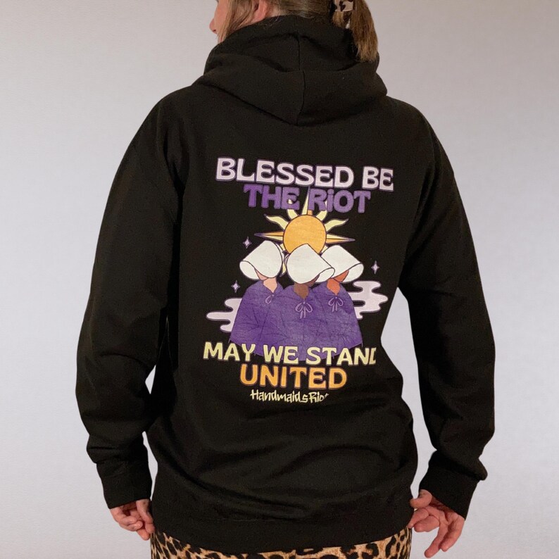 Pode incluir: Moletom preto com a frase "Blessed Be The Riot" e "May We Stand United" em roxo e amarelo. O gr&aacute;fico apresenta tr&ecirc;s figuras com toucas brancas e vestes roxas, com um sol. A marca "Handmaids File" tamb&eacute;m &eacute; vis&iacute;vel.