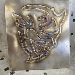 May include: A metal plate with a welded design of a stylized abstract shape. The design is made of multiple layers of welded metal, creating a colorful effect with shades of gold, blue, and purple.