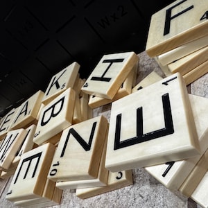 May include: Wooden Scrabble tiles with black letters and numbers. The tiles are stacked and scattered, with letters like 'E', 'A', 'W', and 'B' visible. The background is a dark, textured surface.
