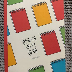 May include: A cream-coloured book with a red spine and a pattern of colourful spiral notebooks. The book title is "Korean Writing Workbook" and the text "JNK Reading Club" is printed below the title.