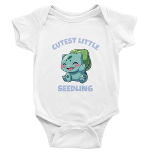 Op de afbeelding: Een wit babyrompertje met korte mouwen en een afbeelding van een groenblauw wezen met een groene bol op de rug. Het wezen heeft roze wangen en zwaait. De tekst erboven luidt CUTEST LITTLE en de tekst eronder SEEDLING in het lichtblauw.