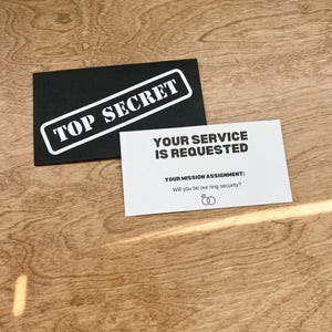 May include: A black card with the words "TOP SECRET" in white letters. Below the card is a white card with the text "YOUR SERVICE IS REQUESTED" and "YOUR MISSION ASSIGNMENT: Will you be our ring security?" with a simple illustration of two interlocking rings.