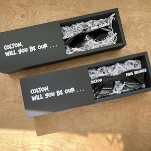 May include: Two black boxes with white text that reads "COLTON, WILL YOU BE OUR..." and "COLTON" on the front. Each box contains a pair of black sunglasses with white text that reads "RING BEARER" and "RING SECURITY".