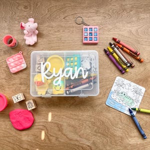 May include: A clear plastic box labeled "Ryan" filled with crayons, playdough, and small toys. Includes a pink elephant, a pink pop-it toy, and a small puzzle with a crocodile illustration. The items are arranged on a wooden surface.