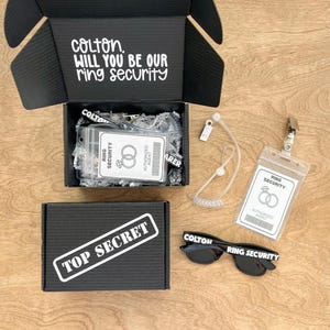 May include: A black box with the text "colton, will you be our ring security" on the top. Inside the box is a black lanyard with a badge that says "ring security" and a pair of black sunglasses with the text "colton" and "ring security" on the arms. The box also has a sticker that says "top secret".