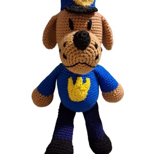 May include: A handmade crochet dog doll in a blue police uniform and hat with a yellow badge. The dog has brown fur, black eyes, and a black nose. The doll is standing upright against a white background.