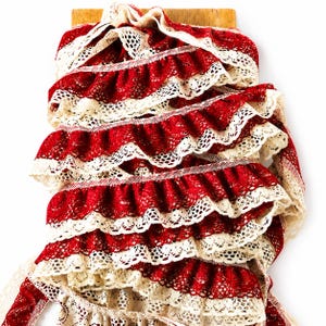 May include: A continuous length of ruffled trim featuring alternating layers of deep red fabric and cream-colored lace. The trim is gathered into multiple horizontal folds, showcasing a repeating pattern of solid red sections bordered by intricate, openwork cream lace edges.
