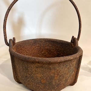 May include: A round, heavy-duty cast iron pot featuring a thick, textured, rusted brown surface. The pot includes a sturdy, arched metal handle attached to two side lugs and rests on three short, integrated feet at the base.