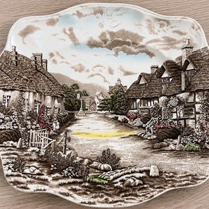 May include: A scalloped ceramic plate featuring a detailed brown and white transferware scene of a village with thatched-roof cottages, a central path, and lush garden foliage. The illustration includes subtle touches of blue and yellow color.