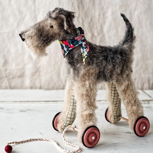 Può includere: Un cane giocattolo grigio e bianco con un bandana a fantasia floreale intorno al collo. Il cane è su ruote e ha una corda attaccata a lui. Il cane è su una superficie di legno bianca.