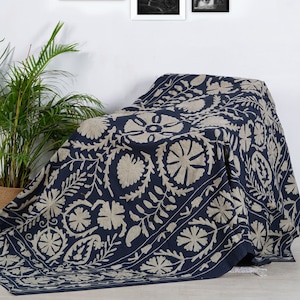 Puede incluir: Un textil bordado en azul marino y beige drapeado sobre una silla, con una colecci&oacute;n de fotograf&iacute;as en blanco y negro enmarcadas en la pared blanca detr&aacute;s. El textil presenta patrones florales y geom&eacute;tricos. Una planta en maceta est&aacute; a la izquierda.