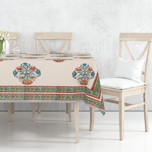 May include: A white tablecloth with a floral pattern in shades of orange, blue, and green. The tablecloth is laid on a wooden table with four chairs. There are white flowers in a vase on the table.