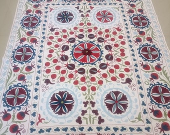 Puede incluir: Una alfombra de algodón blanco con un diseño tradicional bordado. La alfombra presenta un medallón central con un patrón floral y está rodeada por un borde de granadas y motivos florales. Los colores utilizados en el bordado son rojo, azul, verde y marrón.