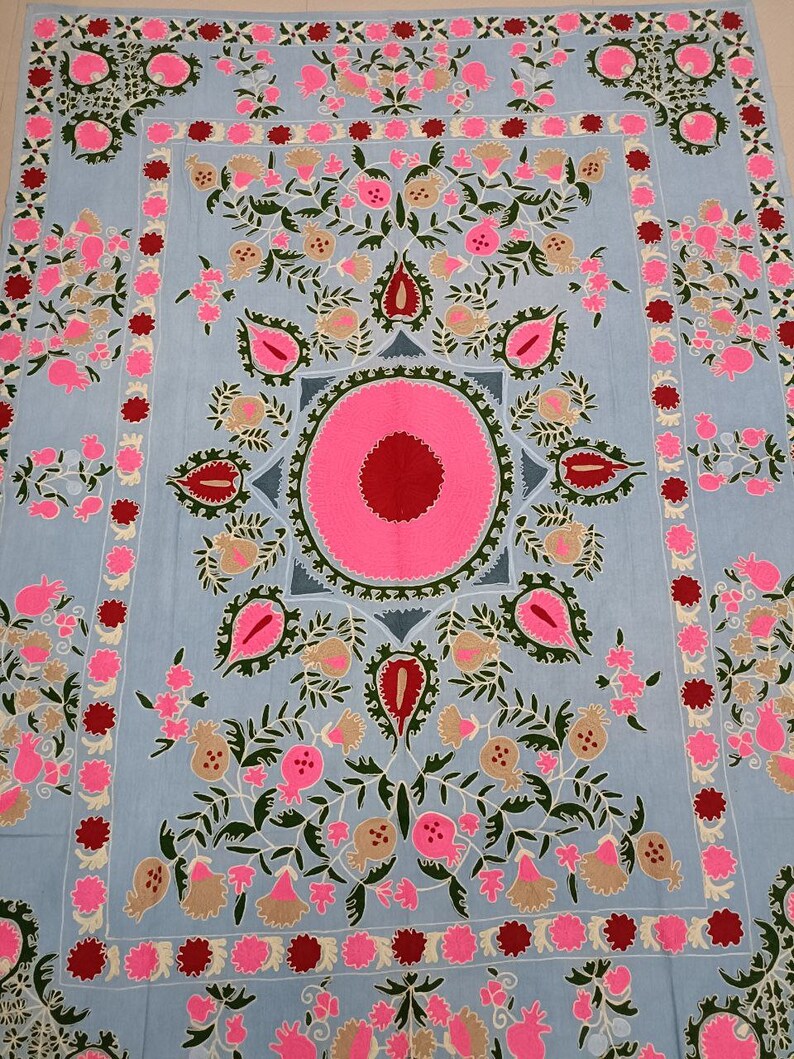 May include: A light blue fabric with intricate embroidery featuring pink, red, and green floral designs. The centre of the fabric features a large pink circle with a red centre.