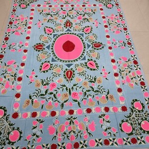 May include: A light blue cotton throw blanket with intricate embroidery featuring pink, green and red floral designs. The centre of the blanket features a large pink circle with a red centre.