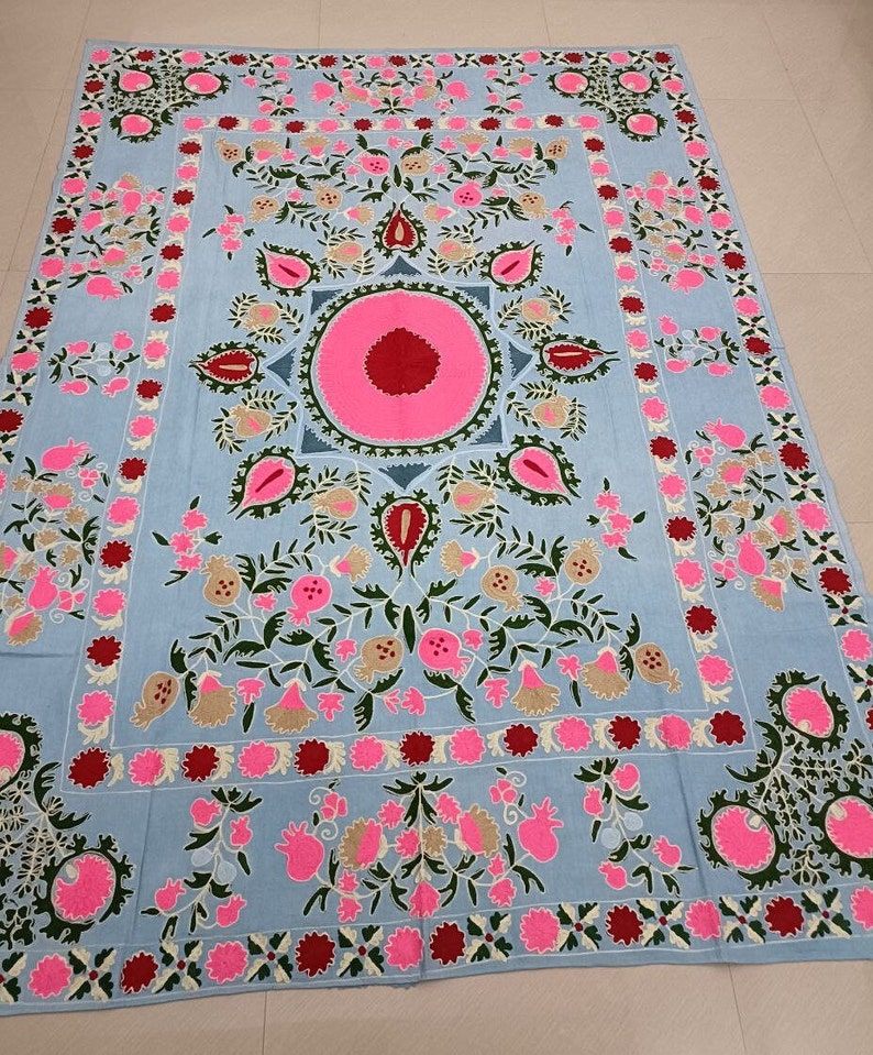 May include: A light blue cotton throw blanket with intricate embroidery featuring pink, green and red floral designs. The centre of the blanket features a large pink circle with a red centre.